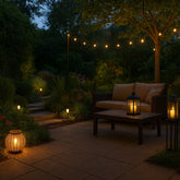 A cozy garden patio at dusk featuring a wicker sofa, wooden table with lanterns, string lights overhead, and pathway lights illuminating stone steps surrounded by lush greenery.